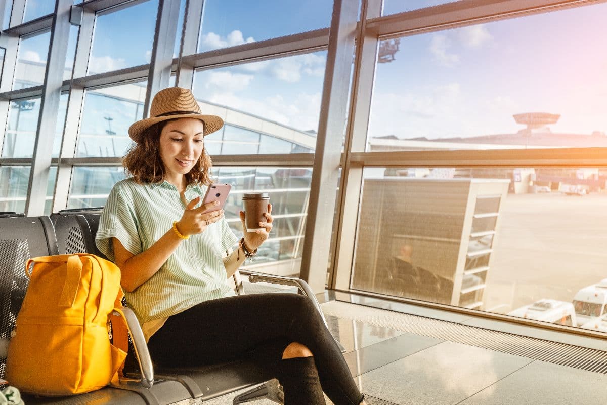 Femme dans salle d'attente d'aéroport pianote sur son smartphone avec le Pass Voyage de Lebara